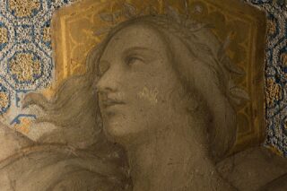 Ein detailliertes Fresko des Gesichts einer Frau mit wallendem Haar und einem Lorbeerkranz vor einem geometrisch gemusterten Hintergrund.