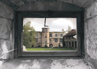Blick durch ein zerbrochenes Fenster auf ein altes, verwittertes Gebäude mit einem grasbewachsenen Innenhof und Bäumen im Hintergrund.
