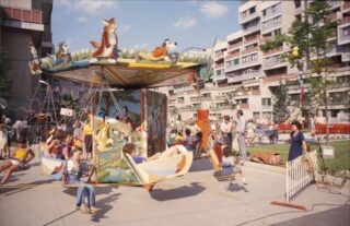 Kinder fahren auf einem Karussell mit Tiermotiven auf einem Jahrmarkt im Freien, während sich Erwachsene und andere Kinder an einem sonnigen Tag in einem städtischen Wohngebiet versammeln.