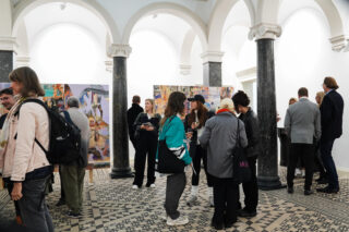 Eine Gruppe von Menschen hat sich in einer Kunstgalerie mit gewölbten Decken und gemusterten Böden versammelt, betrachtet Kunstwerke und unterhält sich.