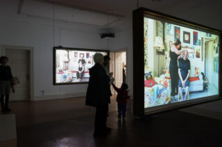 Die Leute schauen auf große Bildschirme, auf denen Fotos einer Frau zu sehen sind, die in einem hell dekorierten Raum in einem schwach beleuchteten Galerieraum einer anderen Frau die Haare frisiert.