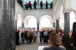 Eine Gruppe von Menschen steht in einer Halle mit Marmorsäulen und gewölbten Decken und hört einem Redner zu; einige stehen auf einem oberen Balkon und im Hintergrund sind farbenfrohe Kunstwerke ausgestellt.