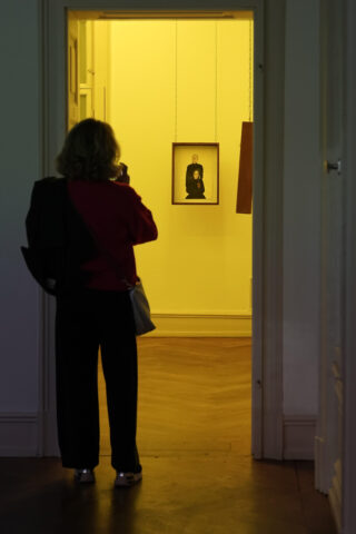 Eine Person steht in einem dunklen Flur und blickt durch eine Tür auf ein gerahmtes Porträt, das an einer gelb beleuchteten Wand hängt.