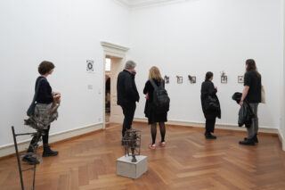 Eine Gruppe von Menschen betrachtet kleine Kunstwerke an einer Galeriewand; auf dem Holzboden des weißwandigen Raums sind Metallskulpturen ausgestellt.