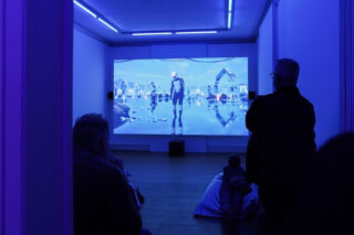 Mehrere Personen sitzen in einem schwach beleuchteten Raum mit blauer Beleuchtung und schauen auf einen großen Bildschirm, auf dem eine futuristische oder digitale Szene angezeigt wird.