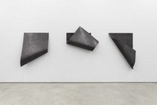 An einer weißen Wand sind drei schwarze, rechteckige Metallplatten montiert, deren Ecken jeweils kunstvoll nach außen gebogen bzw. gefaltet sind. Der Boden besteht aus hellgrauem Beton.