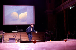 Ein Künstler gestikuliert auf der Bühne mit Orangen auf dem Boden, während dahinter ein projiziertes Bild von zwei Orangen angezeigt wird. Im Hintergrund sind Musikinstrumente zu sehen.