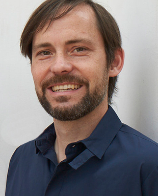 Ein Mann mit kurzen braunen Haaren und Bart, der ein dunkelblaues Hemd mit Kragen trägt, lächelt vor einem schlichten hellen Hintergrund in die Kamera.