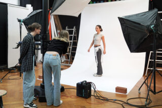 Eine junge Frau posiert vor einem weißen Hintergrund in einem Fotostudio, während zwei Personen in der Nähe einer Kamera und einer Beleuchtungsausrüstung stehen und sich auf ein Fotoshooting vorbereiten.