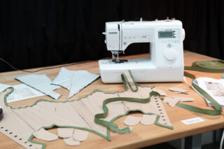 Auf einem Holztisch steht eine Nähmaschine, umgeben von Stoffstücken, Mustern und grünen Besatzmaterialien, wahrscheinlich für die Herstellung von Kleidungsstücken.