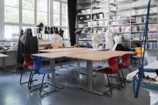 Ein Modedesignstudio mit Schaufensterpuppen, Nähmaschinen, einem großen Holztisch, bunten Stühlen, Regalen mit Zubehör und großen Fenstern, die natürliches Licht hereinlassen.