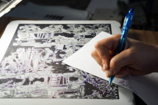 Eine Person macht sich Notizen auf einem kleinen Blatt Papier, während sie eine detaillierte, monochrome Illustration auf einem Tablet oder einer Leuchtbox betrachtet.