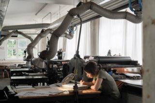 Eine Person sitzt allein an einer Werkbank in einer Werkstatt und konzentriert sich auf das Zeichnen oder Schnitzen unter einer Lampe, umgeben von Geräten, Leitungen und verstreuten Papieren.