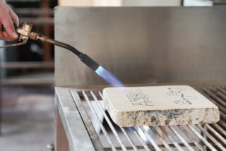 Eine Person erhitzt mit einem Lötbrenner eine Steinplatte mit Zeichnungen, die auf einem Metallgitter liegt.