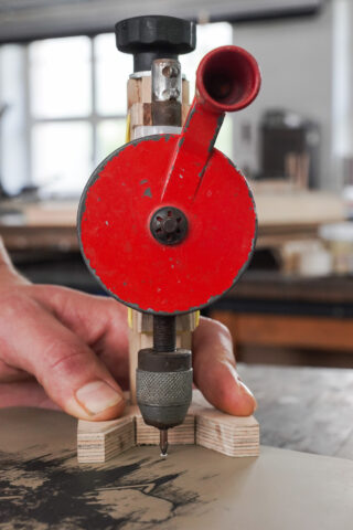 Eine Nahaufnahme einer Hand, die ein rotes mechanisches Gravierwerkzeug auf einem Stück Holz bedient, wobei die Werkzeugspitze die Oberfläche berührt.