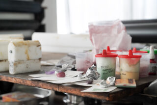 Ein überfüllter Arbeitstisch mit Plastikbehältern, Folie, Bechern, Papier und verschiedenen kleinen Gegenständen, was auf ein laufendes Kunst- oder Wissenschaftsprojekt schließen lässt.