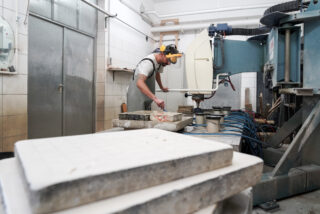 Ein Arbeiter mit Schutzbrille und Gehörschutz bedient Maschinen in einer Industriewerkstatt und bearbeitet eine flache Steinplatte.