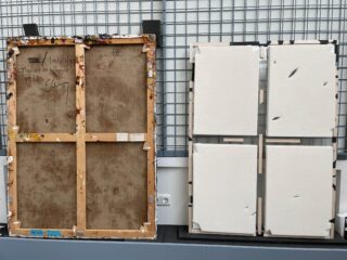 Zwei große Gemälde werden von hinten gezeigt, eines mit Holzkeilrahmen und handschriftlichen Notizen, das andere mit modernem Rahmen und weißer Schutzhülle, beide vor einer Metallgitterwand.