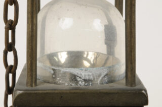 Nahaufnahme einer alten Glaskuppel mit Kondenswasser, die auf einem metallischen Sockel mit daran befestigten Ketten ruht. Im Inneren der Kuppel sind einige weiße Rückstände sichtbar.