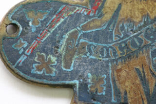 Nahaufnahme eines verwitterten Metallobjekts mit eingravierten und aufgemalten Mustern, darunter blaue und goldene Formen und teilweise Beschriftungen sowie ein Loch in der Nähe der Kante.