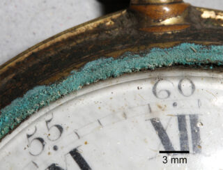 Nahaufnahme einer korrodierten Metallkante auf einem alten Zifferblatt. Sie zeigt grünlich-blaue Korrosion über den Zahlen 55 und 60, mit einer 3-mm-Skala als Referenz.
