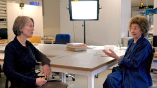 In einem gut beleuchteten Büro oder Studio sitzen sich zwei Frauen an einem Tisch gegenüber und unterhalten sich. Auf dem Tisch liegen Bücher und Papiere.