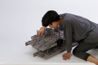 Eine kniende Person richtet ein kleines Architekturmodell mit mehreren Stockwerken und Säulen auf einem schlichten Hintergrund aus.