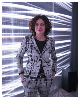 Eine Person mit lockigem Haar und gemustertem Anzug steht vor einem Hintergrund mit horizontalen Lichtstreifen, hat die Hände in den Taschen und lächelt leicht in die Kamera.