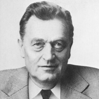 Schwarz-Weiß-Porträt eines älteren Mannes in kariertem Jackett, Hemd mit Kragen und Krawatte, der direkt in die Kamera blickt.