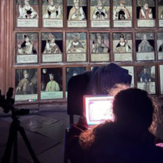 Eine Person sitzt vor einem beleuchteten Laptop und blickt auf eine Wand voller gerahmter Porträts und illustrierter Tafeln. In der Nähe steht ein Stativ.