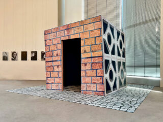 Eine kubische Struktur mit Ziegel- und geometrischen Mustern steht auf einer gefliesten Matte in einer Galerie, an der Wand dahinter hängen Porträts, und rechts davon befinden sich hohe Fensterjalousien.