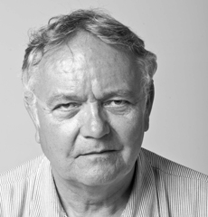 Älterer Mann mit kurzen grauen Haaren und ernstem Gesichtsausdruck, der ein helles, gestreiftes Hemd mit Kragen vor einem schlichten Hintergrund trägt.