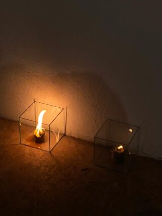 In Glaswürfeln, die auf einer Holzfläche vor einer schlichten Wand stehen, brennen zwei kleine Flammen; eine Flamme ist größer und heller als die andere.