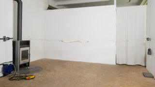 Ein gedrehtes Kabel mit gleichmäßig verteilten weißen Segmenten hängt horizontal zwischen zwei Stützen in einem Raum mit weißen Wänden und einem Holzofen.