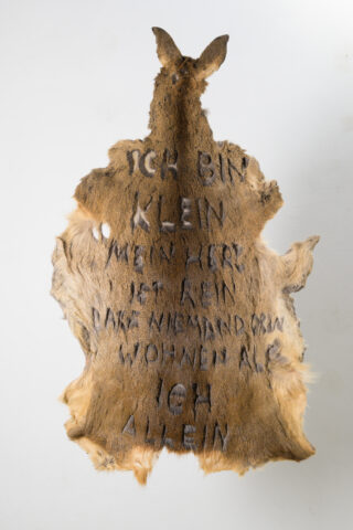 Eine gegerbte Tierhaut mit eingraviertem deutschen Text, flach auf einer weißen Oberfläche ausgestellt. Der Text lautet: „ICH BIN KLEIN UND IM HERZ IST KEIN PLATZ NIEMANDEN WOHNE ICH ALLEIN.