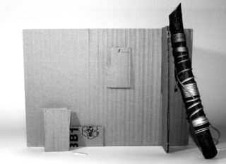 Ein aufrecht stehender Kartonbogen ist mit zwei kleineren Kartonstücken befestigt und ein mit Schnur umwickelter Stock lehnt daran.