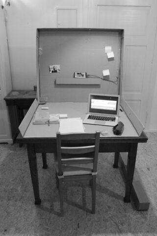 Vor einem Stuhl steht ein Holzschreibtisch mit einem aufgeklappten Laptop, Papieren und Haftnotizen; auf dem Schreibtisch ist ein Kartonhintergrund mit Fotos und Notizen angebracht.