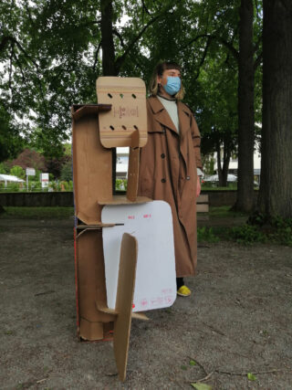 Eine Person in einem braunen Mantel und einer Gesichtsmaske steht im Freien neben einer hohen Pappkonstruktion aus flachgedrückten Kartons und Platten.