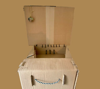 Ein offener Karton mit Deckel, Seilgriff und mehreren Schrauben, die aus der inneren Klappe ragen; an der oberen linken Ecke hängt ein kleines Ornament.