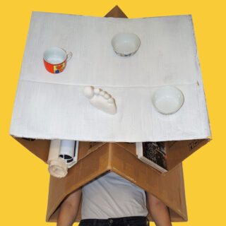 Eine Person unter einem großen Karton mit einer flachen, weiß gestrichenen Oberfläche, die Tassen, Schüsseln und eine weiße Fußskulptur hält, vor einem einfarbig gelben Hintergrund.