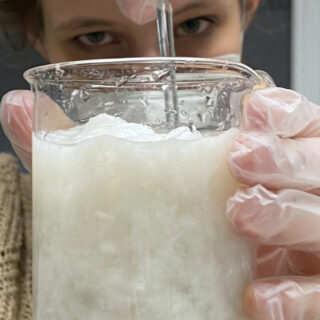 Eine Person mit Handschuhen rührt eine dicke, weiße Substanz in einem Becherglas.