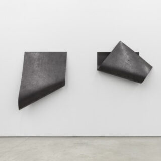 An einer weißen Wand sind zwei schwarze, eckige Metallplatten angebracht, jede mit gebogenen und gefalteten Kanten, wodurch abstrakte, geometrische Formen entstehen.