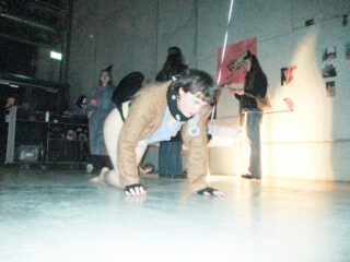 Person in braunem Pullover und Handschuhen kriecht auf Händen und Knien durch einen schwach beleuchteten Raum; mehrere Personen stehen im Hintergrund in der Nähe einer Wand mit Plakaten.