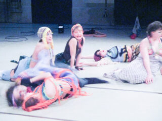 Sechs Menschen in farbenfrohen, vielseitigen Kostümen liegen und sitzen im Innenbereich auf dem Boden, einige blicken in die Kamera, wobei die Bewegungsunschärfe eine Bewegung suggeriert.