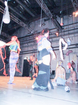 Menschen in farbenfrohen, vielseitigen Kostümen treten auf oder interagieren auf einer Bühne mit Deckenbeleuchtung und verschiedenen hängenden Requisiten in einem Ambiente im Industriestil.