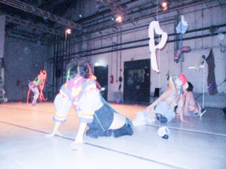 Drei Personen in farbenfrohen, vielseitigen Kostümen treten auf einer schwach beleuchteten Bühne mit abstrakten hängenden Objekten und industriellen Hintergrundelementen auf.