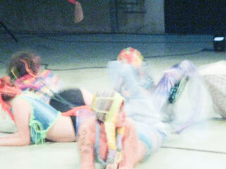 Drei Menschen in farbenfrohen Kostümen und mit Kopfbedeckungen aus Garn liegen in einer verschwommenen, bewegungsreichen Szene im Innenbereich auf dem Boden.