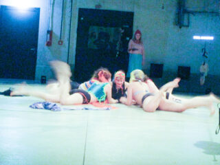 Vier Personen in bunten Kostümen liegen in einem beleuchteten Innenraum auf dem Boden, während eine andere Person im Hintergrund steht und sie beobachtet.