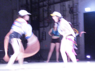 Drei Personen stehen im Innenbereich. Zwei im Vordergrund scheinen sich schnell zu bewegen, was zu Unschärfe führt. Sie tragen Hüte und bunte Kleidung. Eine dritte Person steht im Hintergrund.