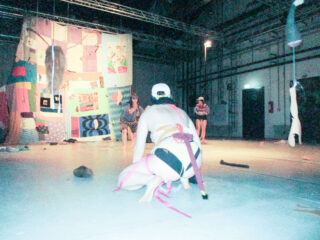 Drei Personen treten auf einer Bühne auf, auf der Patchworkdecken und Gegenstände hängen, während eine Person im Vordergrund kniet und nur minimale Kleidung und rosa Träger trägt.